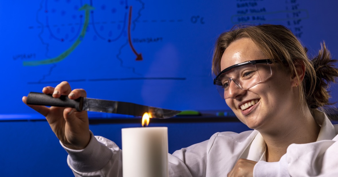 Anika Jane Beamer smiles as she performs a lab experiment by holding a small kitchen knife over a lit candle. She wears a white lab coat and protective glasses