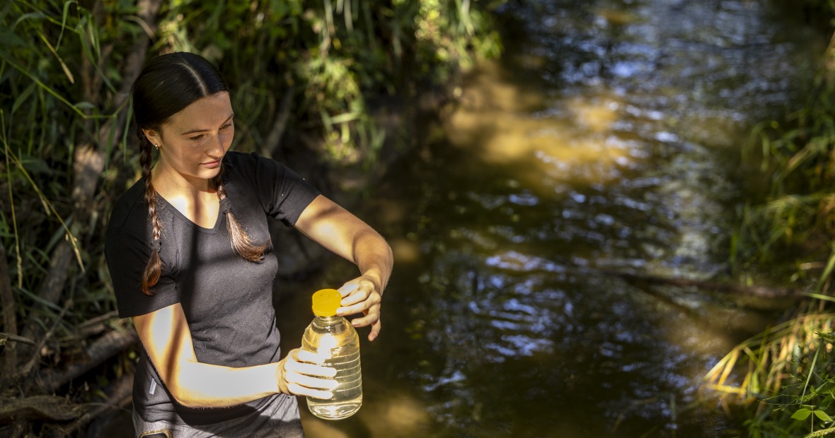 Student Millie Leonard stands in a small creek, holding a large, clear bottle containing a water sample.