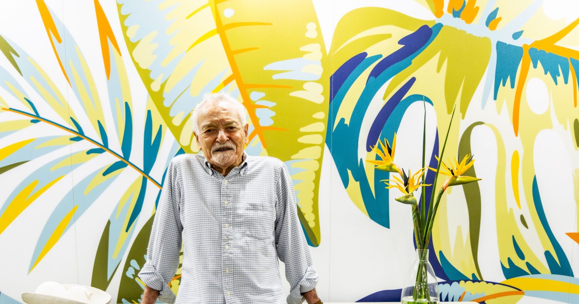 Tony Dalsimer, a senior person, wearing a striped button-up shirt leans on a white surface with a colorful tropical leaf mural and a vase with bird of paradise flowers in the background. 