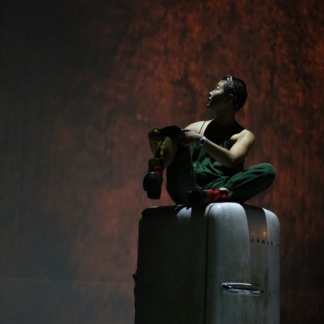 A performer in overalls perches atop an aged vintage refrigerator under dramatic light