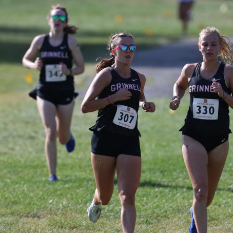 Two female college students wearing cross country uniforms run together, a third behind them