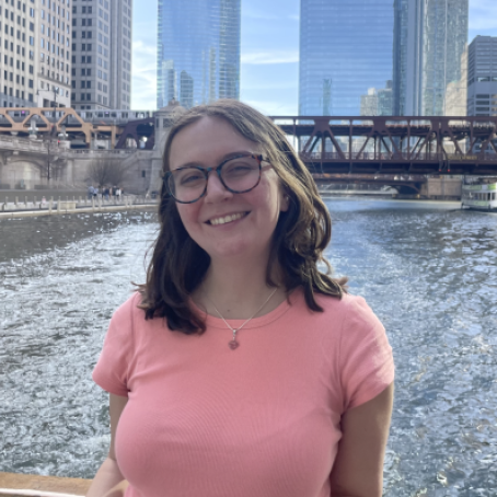 Claire Giannosa, a brunette woman with glasses, wears a pink shirt. She stands on a bridge overlooking a river in a city