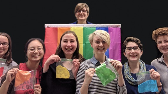 Members of Out in STEM holding bags of a colored substance to match the pride flag behind them
