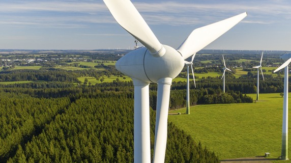 Large wind turbine is first in a series of them with a green field and forest in the background