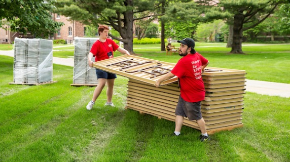 Two students lift a folding table from a stack of them to help set up for reunion