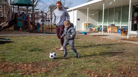child plays soccer with caregiver at preschool playground