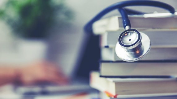 Focused shot of stethoscope draped over books with unfocused hand typing on laptop in background