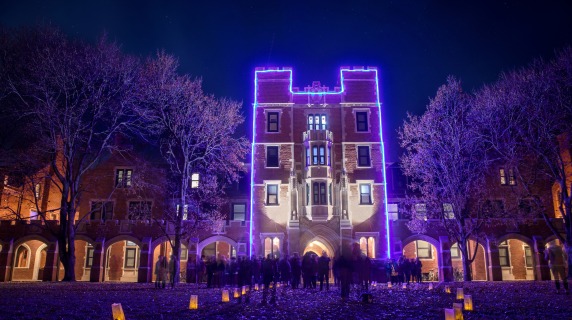 Gates Hall at night in the winter. The building is outlined in blue lights with a trail of paper bag candles leading up to it.