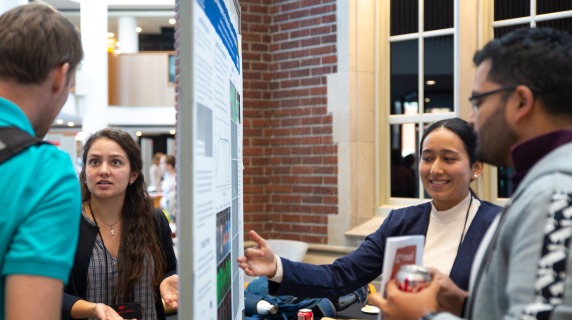 Two women present their research posters in a busy atrium. They stand on either side of a bulletin board, and are gesticulating toward their posters while speaking to listeners in the foreground.