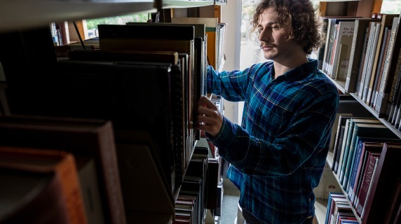 A young man stands in the library stacks examining a book