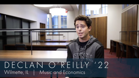 Declan O'Reilly '22, Music and Economics major from Wilmette, IL sits in a reading room in the humanities building on campus. He faces the camera. There are many tables with reading lamps behind him.