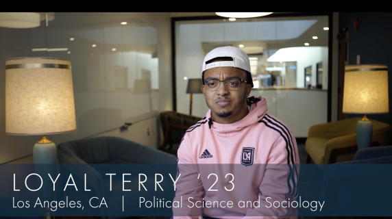 Loyal Terry '23, a Political Science and Sociology major from Los Angeles, CA, sits in a student study space in the humanities building. There are lounge chairs, well-lit lamps, and a glass window behind him that reveals the building hallways.