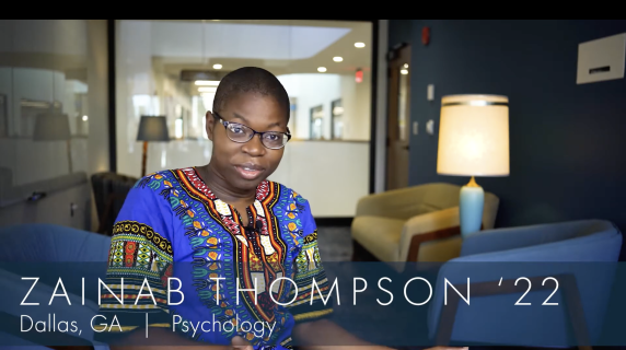 Zainab Thompson '22, Psychology major from Dallas, GA, sits in one of the student study spaces in the humanities building. Behind them are lounge chairs, well-lit lamps, and a glass window that reveals the hallways of the humanities building.