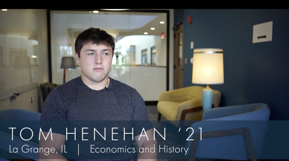 Tom Henehan '21, Economics and History major from La Grange, IL, sits in one of the study spaces in the humanities building. Behind him are lounge chairs, well-lit lamps, and a glass window that reveals the hallways of the humanities building.
