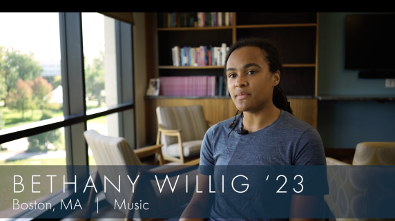 Bethany Willig '23, Music major from Boston, MA looks at the camera and sits in a well-lit student study space with wide open windows. Behind them are bookshelves and chairs that face each other.