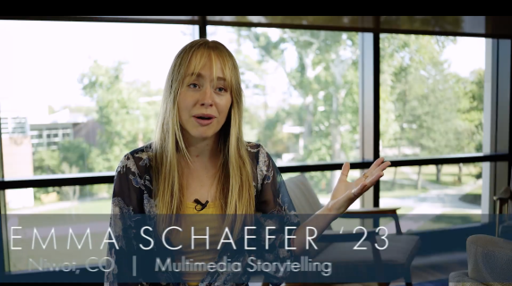 Emma Schaefer '23, Multimedia Storytelling Independent Major from Niwot, CO, sits in a well-lit room and faces the camera as her hand moves with her words. To the back of her are wide windows that reveal a green and bright campus.