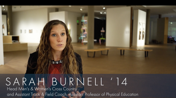 Sarah Burnell '14, Head Men's and Women's Cross Country and Assistant Professor of Physical Education, looks at the camera. Behind her are multiple art displays from Grinnell's Museum of Art.