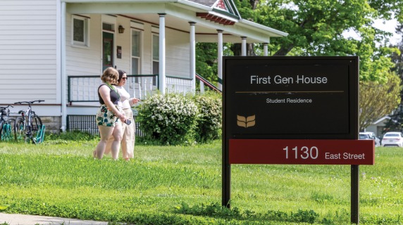A large sign in front of a house reads in white letters on black First Gen House Student Residence the address, 1103 East, appears in white text on a red bar below. 2 women stroll by in the background
