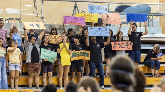 international college students standing in a gymnasium hold up signs in various languages