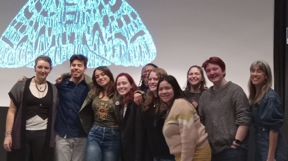 Slam participants and producers stand on stage with a white moth logo in the background.