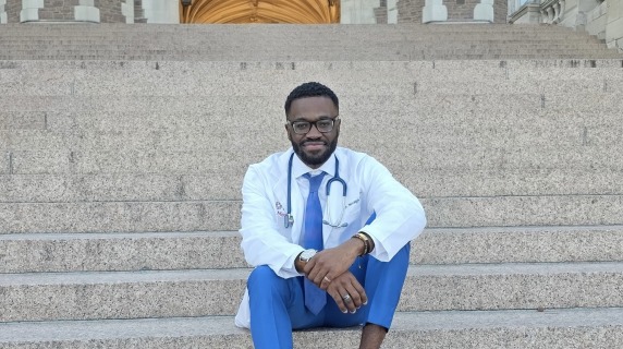 MJ Ketcha wearing a white medical coat and sitting on stone steps.