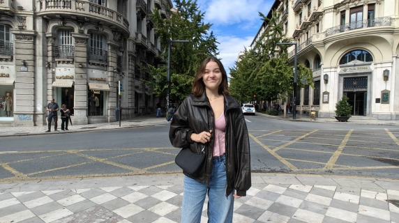 woman with dark hair wearing dark jacket and blue jeans standing in front of buildings