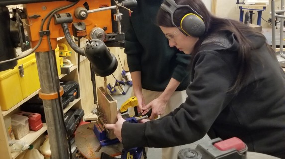 Students wearing safety gear use the drill press in the woodshop.