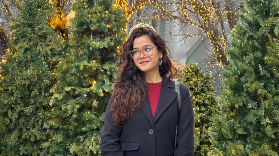 Meghna stands in front of a number of trees, some lit with white holiday lights
