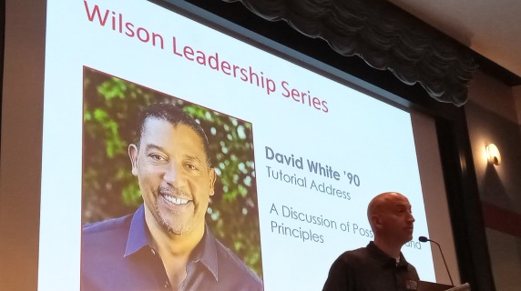 A man at podium to introduce a guest speaker, with intro slide in background. 