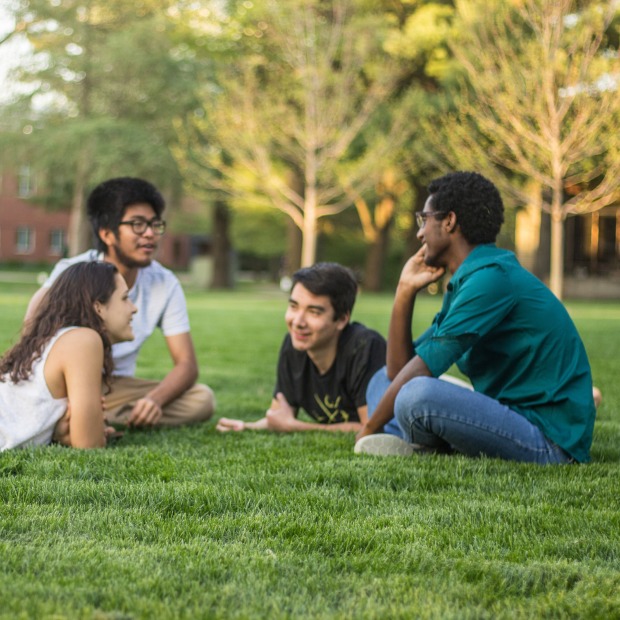 4 students face each other in a circle as they sit or lie on the grass