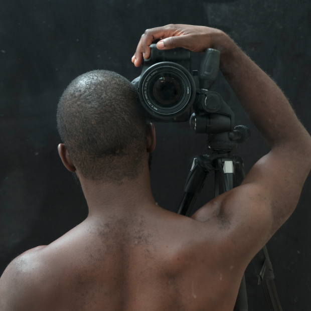 View of a Black figure from behind with a camera perched on his shoulder aimed at the viewer.