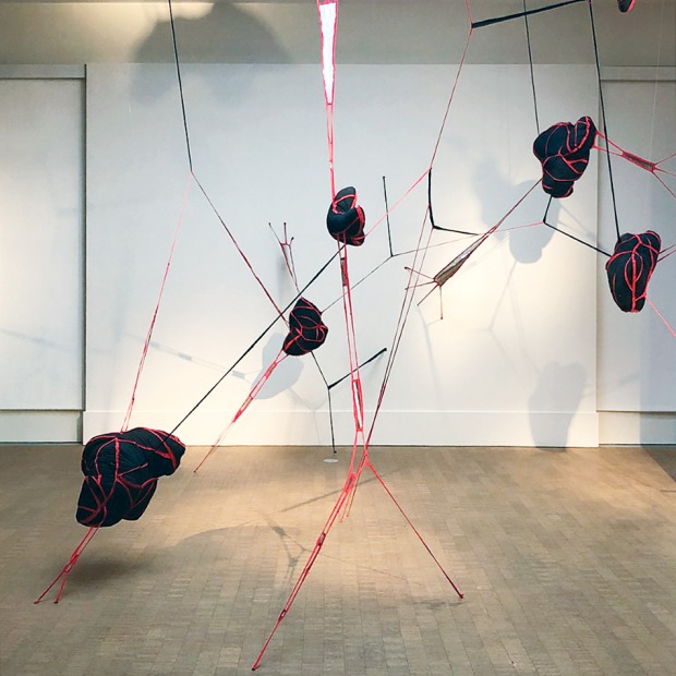Black forms suspended from the walls and ceiling in the gallery by red bands