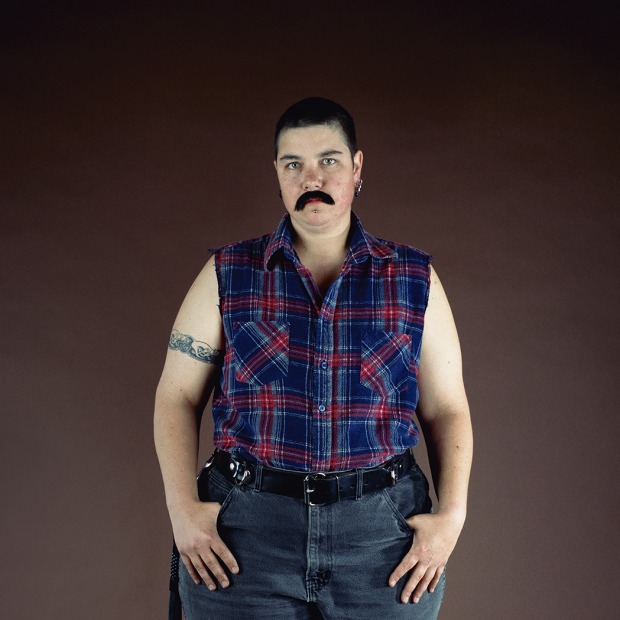 Full-length frontal portrait of the artist Catherine Opie wearing jeans, a sleeveless flannel shirt, and a fake black mustache.