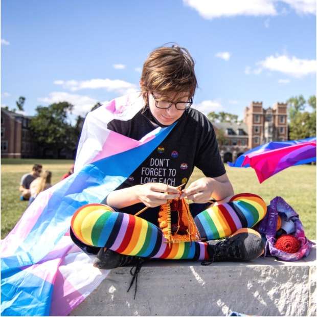 Student with rainbow socks and a trans flag cape sitting outside in the sun knitting