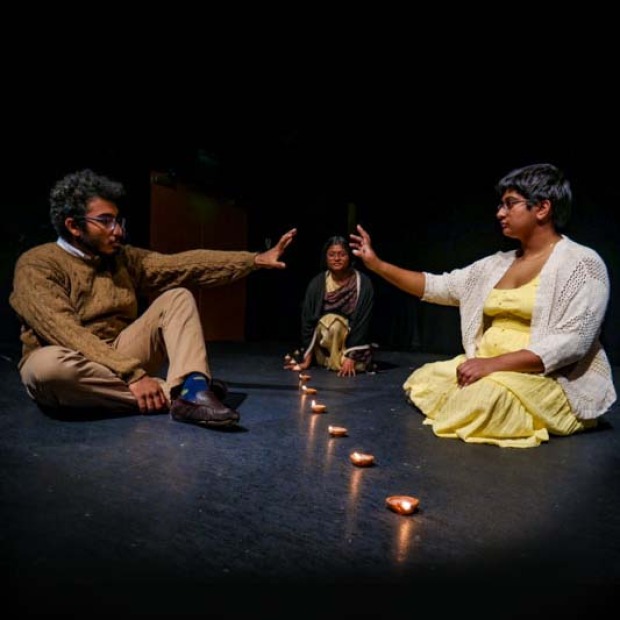 Performers reach toward each other across a line of tea lights in the Wall Studio Theatre
