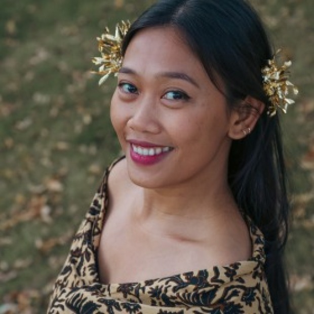 Dewa: an easy smile, long black hair, patterned top, festive gold hair ornaments, and a grassy background.