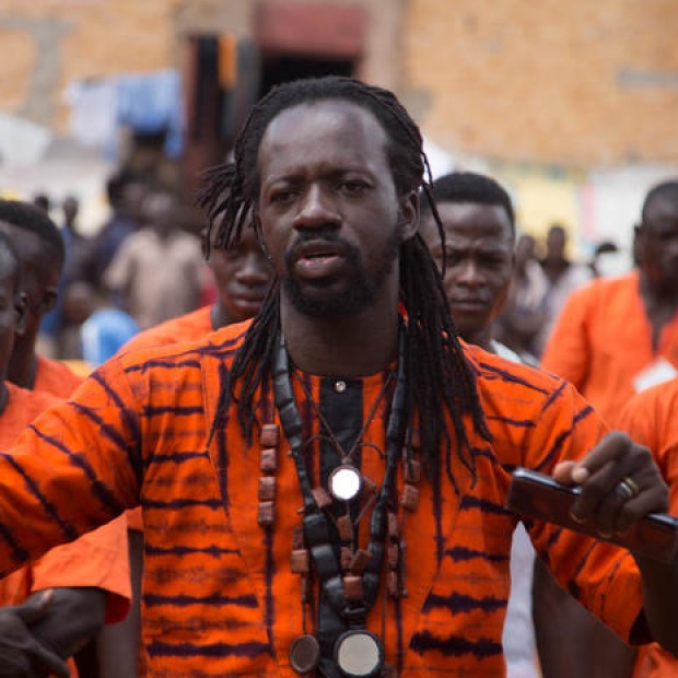 Aguibou: A Burkinabe man with long hair and a vibrant orange tunic gives instruction to a group of incarcerated performers