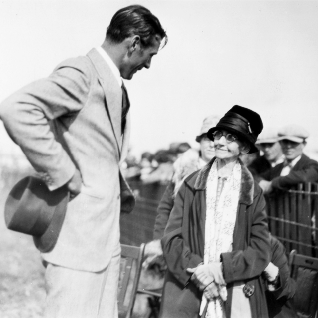 Gary Cooper, arms akimbo, speaking to an older woman, with others behind a temporary fence in the background