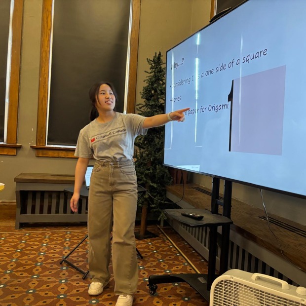 Momoka stands in front of a large screen and points at a large square on it. The screen also includes words explaining an origami concept.