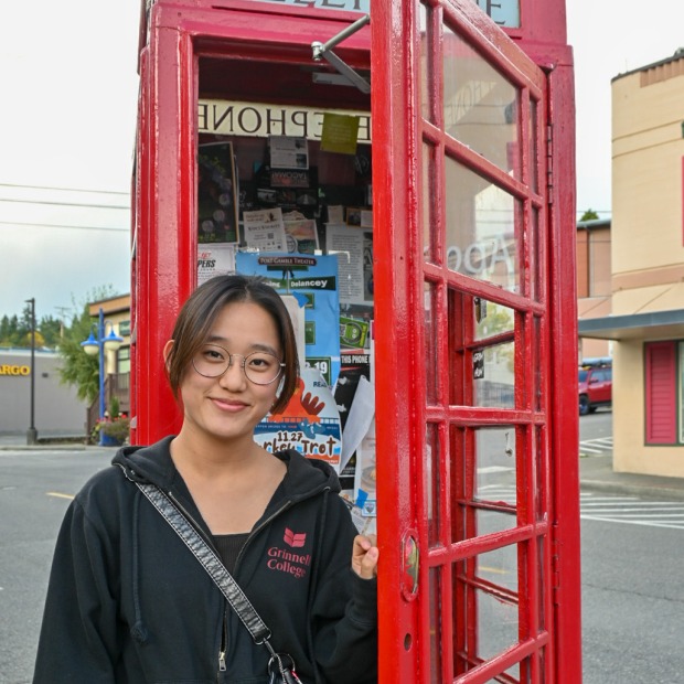Hana smiles at the camera standing in a red telephone booth