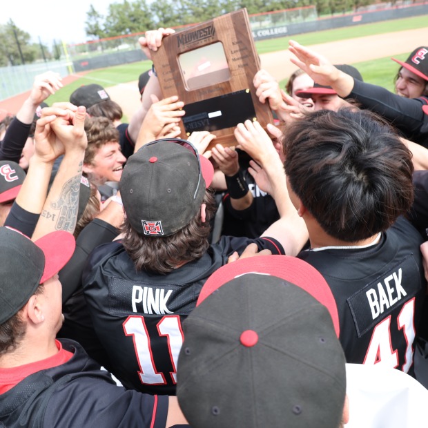 Baseball players wearing black uniforms with red accents celebrate in a group holding a wooden plaque