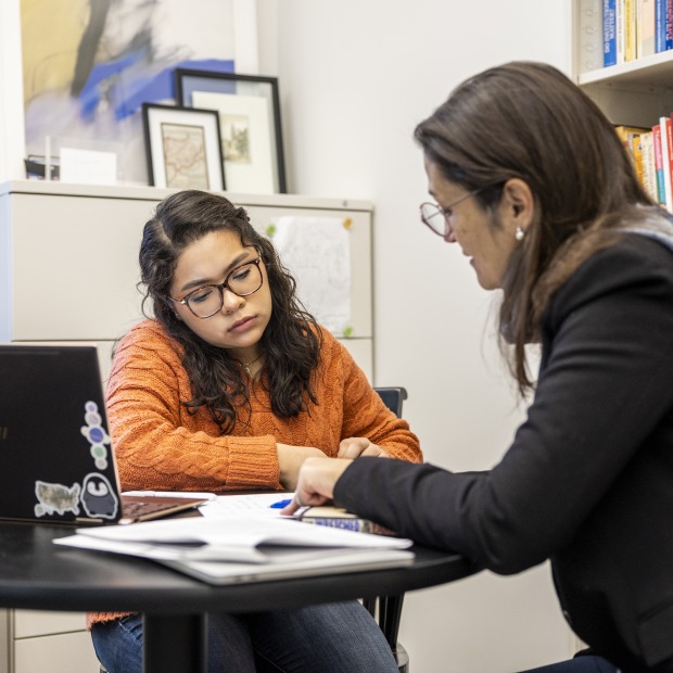 A professor and student sit at a table in an office. The professor points at something on a sheet of paper between them.