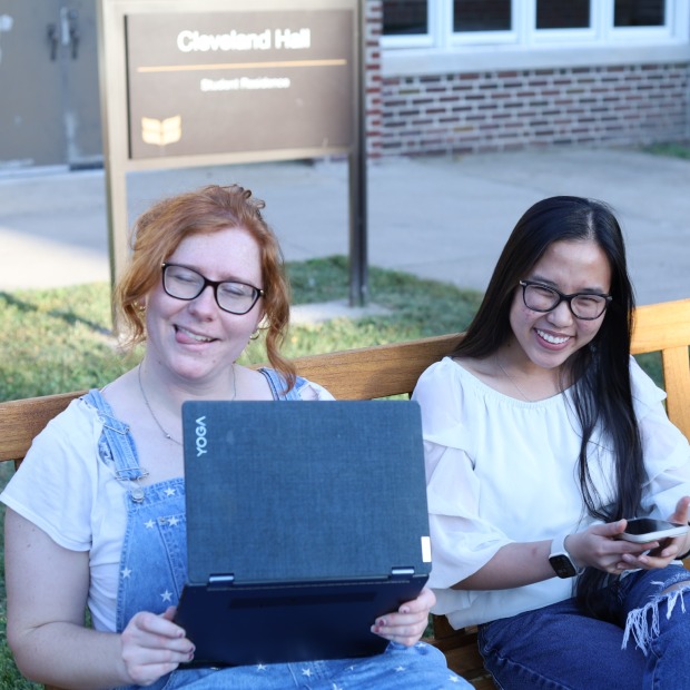 two students sit on a bench, one smiling and the other sticking her tongue out while working on her laptop