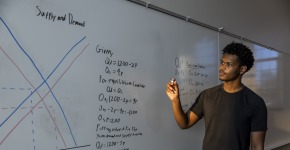 A young man stands next to a white board with the words "Supply and Demand" and a chart and equations