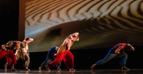 A collections of student performers dance in unison onstage in front of a large projected image of a sand dune. The students wear flowing apparel in jewel tones, and the dance is dramatically lit with a modern feel.