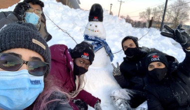 A group of friends in heavy winter coats and ski masks builds a snowman.