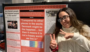 A smiling young woman poses with an academic poster about life in Denmark