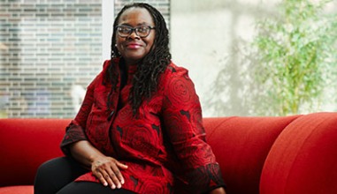 Onwuachi-Willig, in a bright red silky blazer, sits on a red couch in front of a glass window background.