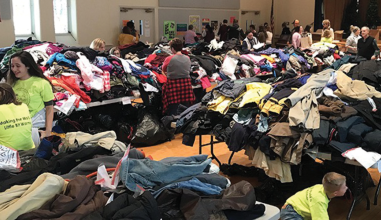 tables overflowing with winter clothing and three people in matching tshirts that read making the world a little bit warmer