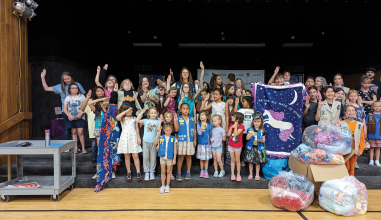 Large group of girls saluting. A woman holds up a blanket with a unicorn against a starry sky that reads we are all made of stars. To the right are several sacks of blankets